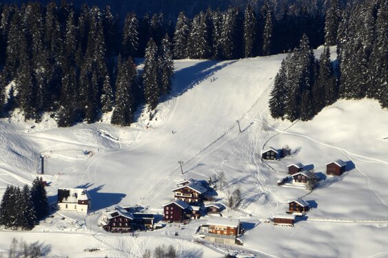 Aerial view of the perfectly groomed ski slope at Kristberg in Silbertal – the mountain of indulgence in the Montafon – with the snow-covered St. Agatha Church and Panoramagasthof Kristberg in the Austrian Alps.
