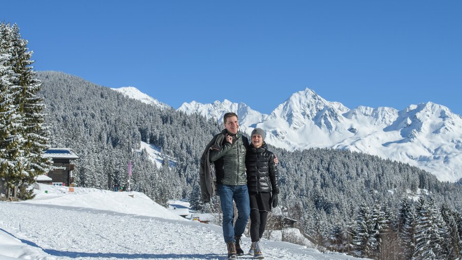Glückliches Paar beim Winterwandern am Kristberg im Silbertal an einem sonnigen Tag, umgeben von tief verschneiter Landschaft und den weißen Gipfeln von Muttjöchle, Lobspitze und Lobschild im Verwallgebirge der österreichischen Alpen. | © Montafoner Kristbergbahn Silbertal GmbH - Patrick Säly Photography - https://patricksaely.com