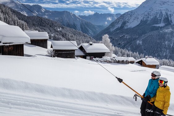 Two female skiers on a T-bar lift in a deeply snow-covered winter landscape with panoramic views of the Silbertal Lobspitz massif in the Austrian Alps.