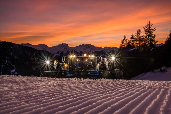 Magical winter moment at Kristbergsattel: A snow groomer in front of a breathtaking sunset, with views of the Drei Türme and the Sulzfluh in the Rätikon mountains of the Austrian Alps – the slope freshly and perfectly groomed.